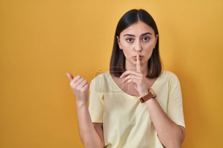 Photo for Hispanic girl wearing casual t shirt over yellow background asking to be quiet with finger on lips pointing with hand to the side. silence and secret concept. - Royalty Free Image