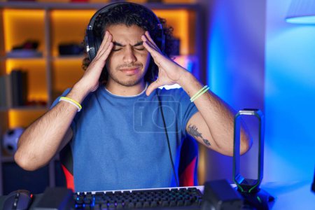 Téléchargez les photos : Homme hispanique avec les cheveux bouclés jouer à des jeux vidéo avec la main sur la tête, maux de tête parce que le stress. migraine souffrant. - en image libre de droit