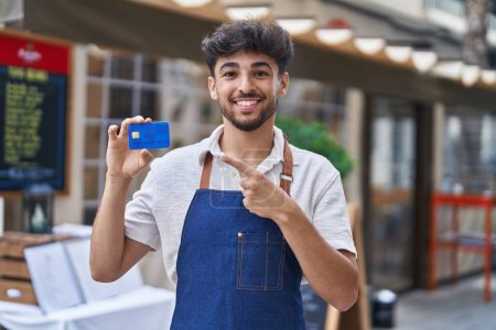 Téléchargez les photos : Arabe avec barbe portant tablier de serveur à la terrasse du restaurant tenant carte de crédit souriant heureux pointant avec la main et le doigt - en image libre de droit