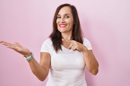Téléchargez les photos : Moyen âge brunette femme debout sur fond rose étonné et souriant à la caméra tout en présentant avec la main et pointant du doigt. - en image libre de droit