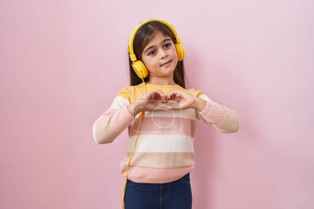 Little hispanic girl listening to music using headphones smiling in love doing heart symbol shape with hands. romantic concept.