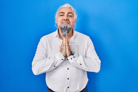 Photo for Middle age man with grey hair standing over blue background praying with hands together asking for forgiveness smiling confident. - Royalty Free Image