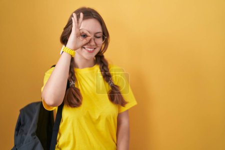 Foto de Joven mujer caucásica con mochila de estudiante sobre fondo amarillo haciendo un gesto bien con la mano sonriente, ojos mirando a través de los dedos con cara feliz. - Imagen libre de derechos