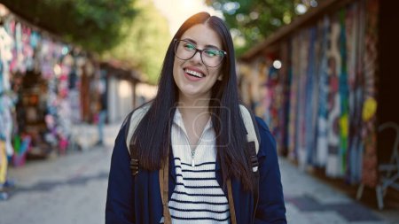 Téléchargez les photos : Jeune hispanique femme touriste portant sac à dos souriant au marché de la rue - en image libre de droit