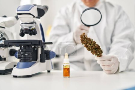 Téléchargez les photos : Jeune homme latin scientifique regardant herbe de cannabis avec loupe au laboratoire - en image libre de droit
