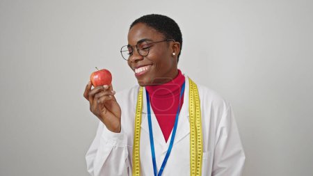 Téléchargez les photos : Femme afro-américaine diététicienne souriante confiante tenant pomme sur fond blanc isolé - en image libre de droit