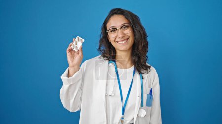 Téléchargez les photos : Jeune belle femme hispanique médecin tenant des pilules sur fond de mur bleu isolé - en image libre de droit