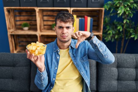Photo for Young hispanic man holding potato chips with angry face, negative sign showing dislike with thumbs down, rejection concept - Royalty Free Image