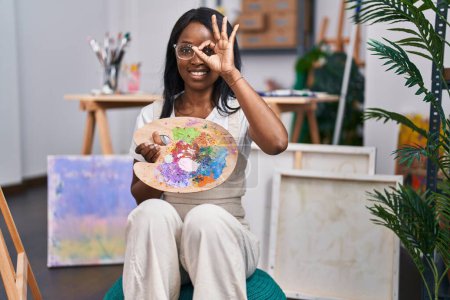 Foto de Mujer joven africana sosteniendo paleta de pintor sonriendo feliz haciendo signo de ok con la mano en el ojo mirando a través de los dedos - Imagen libre de derechos