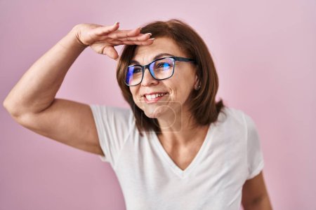 Photo for Middle age hispanic woman standing over pink background very happy and smiling looking far away with hand over head. searching concept. - Royalty Free Image
