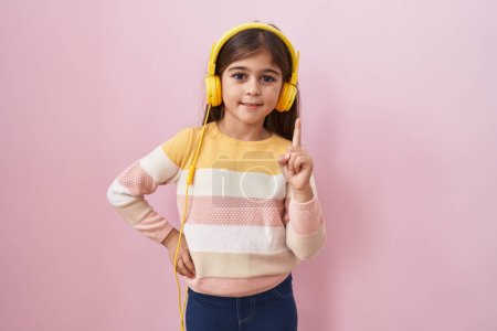 Téléchargez les photos : Petite fille hispanique écoutant de la musique à l'aide d'écouteurs souriant avec une idée ou une question pointant du doigt avec un visage heureux, numéro un - en image libre de droit