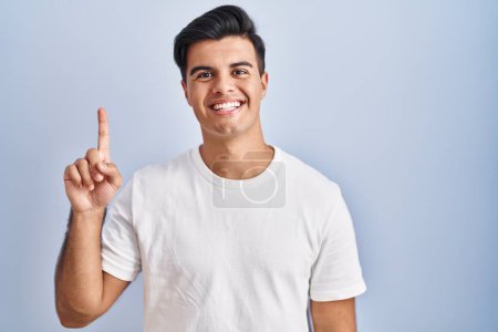 Téléchargez les photos : Homme hispanique debout sur fond bleu montrant et pointant vers le haut avec le doigt numéro un tout en souriant confiant et heureux. - en image libre de droit
