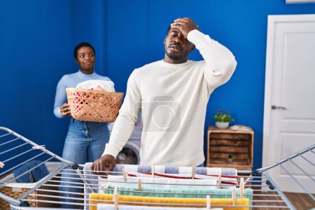 Foto de Joven pareja afroamericana colgando ropa en tendedero estresado y frustrado con la mano en la cabeza, cara sorprendida y enojada - Imagen libre de derechos