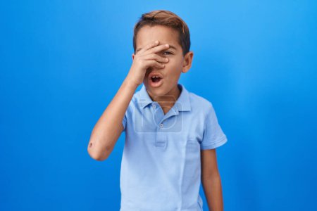 Foto de Pequeño niño hispano vistiendo una camiseta azul casual asomándose en shock cubriendo cara y ojos con la mano, mirando a través de los dedos asustado - Imagen libre de derechos