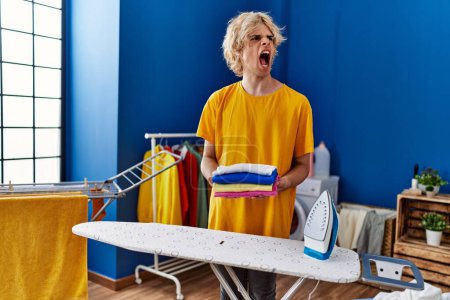 Photo for Young man ironing clothes at home angry and mad screaming frustrated and furious, shouting with anger. rage and aggressive concept. - Royalty Free Image