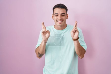 Téléchargez les photos : Bel homme hispanique debout sur fond rose doigt gestuel croisé souriant avec espoir et les yeux fermés. chance et concept superstitieux. - en image libre de droit
