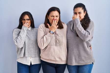 Photo for Mother and two daughters standing over blue background smelling something stinky and disgusting, intolerable smell, holding breath with fingers on nose. bad smell - Royalty Free Image