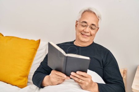 Photo for Senior man reading book sitting on bed at bedroom - Royalty Free Image