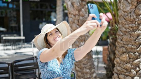 Foto de Joven mujer rubia turista sonriendo confiado hacer selfie por teléfono inteligente en la terraza de la cafetería - Imagen libre de derechos