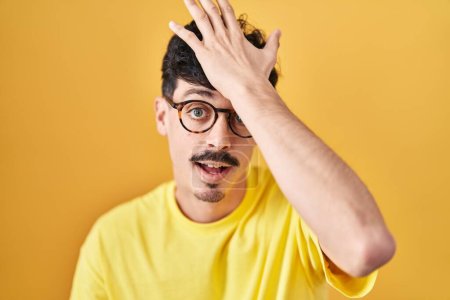 Photo for Hispanic man wearing glasses standing over yellow background surprised with hand on head for mistake, remember error. forgot, bad memory concept. - Royalty Free Image