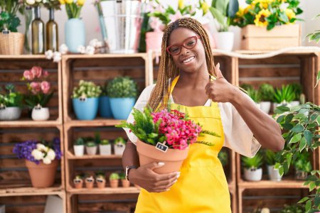 Foto de Mujer afroamericana con cabello trenzado trabajando en floristería sosteniendo planta sonriendo feliz y positiva, pulgar hacia arriba haciendo excelente y signo de aprobación - Imagen libre de derechos