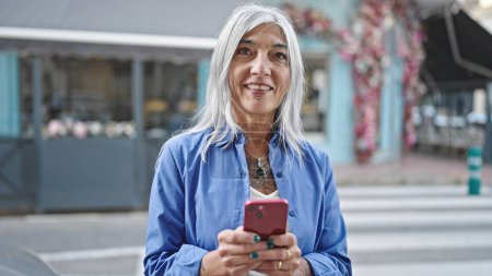 Téléchargez les photos : Femme aux cheveux gris du Moyen Âge utilisant un smartphone souriant sur la terrasse du café - en image libre de droit