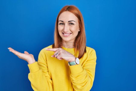 Foto de Mujer joven de pie sobre fondo azul asombrado y sonriendo a la cámara mientras se presenta con la mano y señalando con el dedo. - Imagen libre de derechos