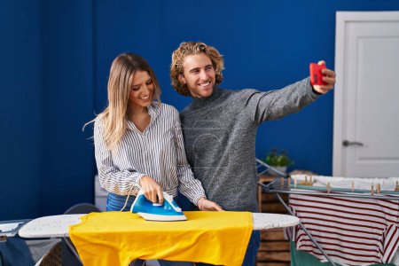 Foto de Hombre y mujer pareja hacer selfie por el teléfono inteligente planchado de ropa en la sala de lavandería - Imagen libre de derechos
