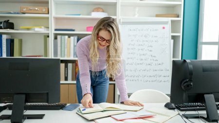 Photo for Young blonde woman teacher smiling confident writing on notebook at classroom - Royalty Free Image