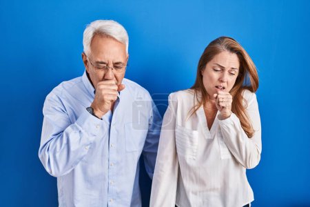 Téléchargez les photos : Couple hispanique du moyen âge debout sur fond bleu se sentant mal à l'aise et toussant comme symptôme de rhume ou de bronchite. concept de soins de santé. - en image libre de droit