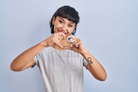 Téléchargez les photos : Jeune femme hispanique portant t-shirt décontracté sur fond bleu souriant dans l'amour faisant forme de symbole de coeur avec les mains. concept romantique. - en image libre de droit