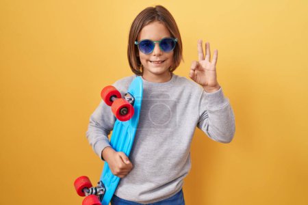 Foto de Pequeño niño hispano con gafas de sol sosteniendo patín haciendo signo de ok con los dedos, sonriendo gesto amistoso excelente símbolo - Imagen libre de derechos