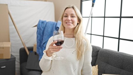 Foto de Joven mujer rubia bebiendo copa de vino sentado en el sofá en el nuevo hogar - Imagen libre de derechos