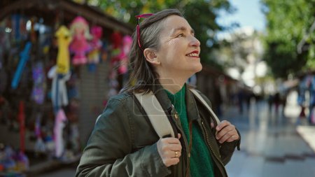 Téléchargez les photos : Femme hispanique mature avec des cheveux gris touriste portant un sac à dos souriant regardant sur le côté au marché de rue - en image libre de droit