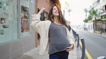 Foto de Joven mujer embarazada sonriendo confiada haciendo un gesto fuerte con el brazo en la calle - Imagen libre de derechos