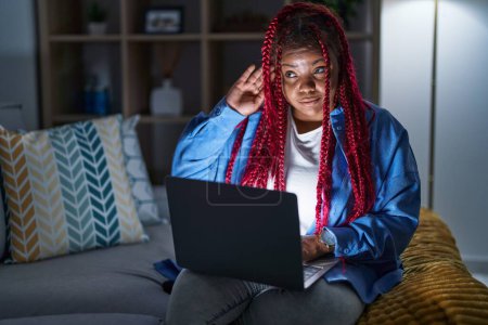 Photo for African american woman with braided hair using computer laptop at night smiling with hand over ear listening an hearing to rumor or gossip. deafness concept. - Royalty Free Image
