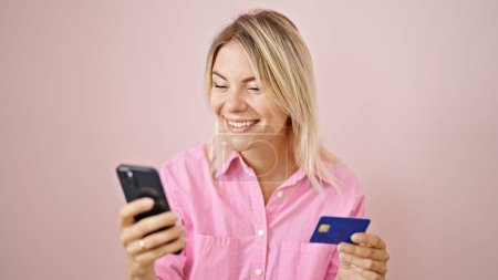 Téléchargez les photos : Jeune femme blonde faisant du shopping en ligne avec smartphone et carte de crédit sur fond rose isolé - en image libre de droit