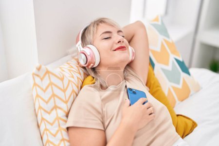 Young blonde woman listening to music relaxed on bed at bedroom