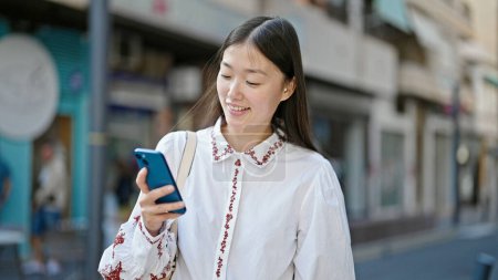 Photo for Young chinese woman using smartphone smiling at street - Royalty Free Image