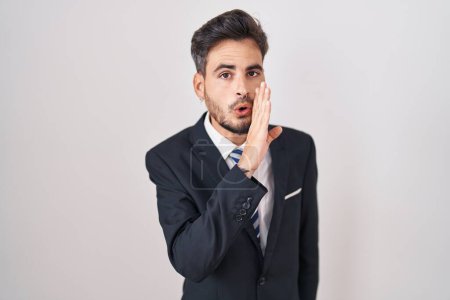 Téléchargez les photos : Jeune homme hispanique avec des tatouages portant un costume d'affaires et une cravate main sur bouche racontant une rumeur secrète, chuchotant une conversation malicieuse - en image libre de droit