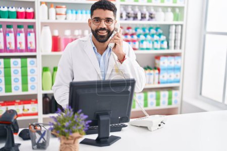 Photo for Young arab man pharmacist talking on telephone using computer at pharmacy - Royalty Free Image