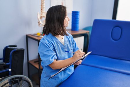Téléchargez les photos : Femme trisomique portant un uniforme de physiothérapie écrivant sur presse-papiers à la clinique de physiothérapeute - en image libre de droit