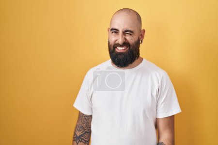 Foto de Joven hombre hispano con barba y tatuajes de pie sobre fondo amarillo guiñando el ojo mirando a la cámara con expresión sexy, cara alegre y feliz. - Imagen libre de derechos