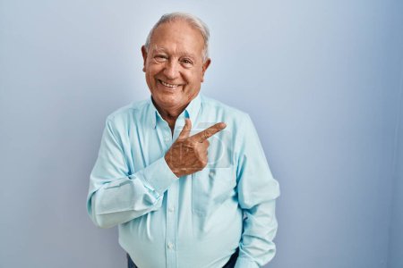 Foto de Hombre mayor con el pelo gris de pie sobre fondo azul alegre con una sonrisa de la cara señalando con la mano y el dedo hacia arriba a un lado con expresión feliz y natural en la cara - Imagen libre de derechos