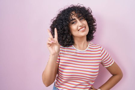 Foto de Joven mujer de Oriente Medio de pie sobre fondo rosa sonriendo mirando a la cámara mostrando los dedos haciendo signo de victoria. número dos. - Imagen libre de derechos