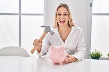 Photo for Young blonde woman holding piggy bank and hammer smiling and laughing hard out loud because funny crazy joke. - Royalty Free Image