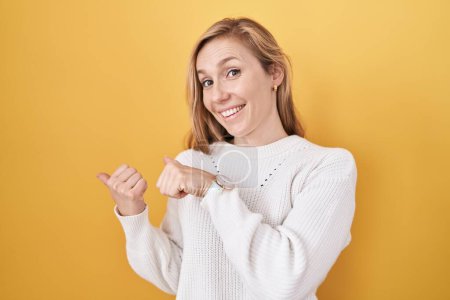 Téléchargez les photos : Jeune femme caucasienne portant un pull blanc sur fond jaune pointant vers le dos avec la main et les pouces vers le haut, souriant confiant - en image libre de droit