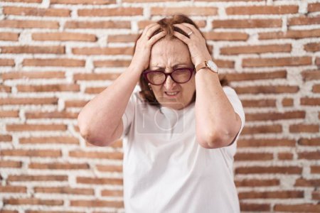 Photo for Senior woman with glasses standing over bricks wall suffering from headache desperate and stressed because pain and migraine. hands on head. - Royalty Free Image