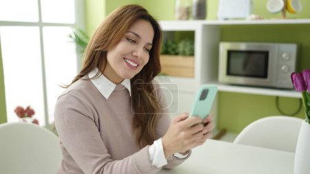 Photo for Young beautiful hispanic woman using smartphone sitting on table at home - Royalty Free Image