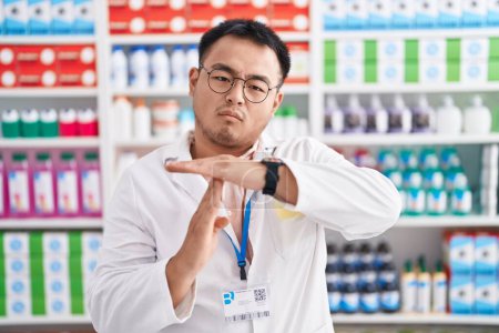 Foto de Joven chino trabajando en farmacia haciendo tiempo fuera gesto con las manos, rostro frustrado y serio - Imagen libre de derechos